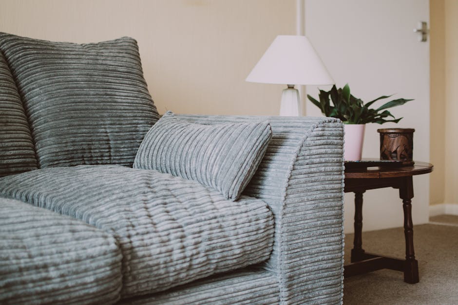 Close-up of a person's hand using a vacuum cleaner with a black nozzle attachment to deep clean an olive-green upholstered armchair. In the background, there is a dark gray fabric sofa, and the room i